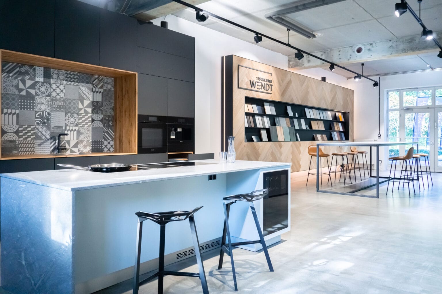 Projekt: Showroom Tischlerei Wendt – Kochinsel mit Barhockern vor dunkler Küchenzeile und Fliesenwand